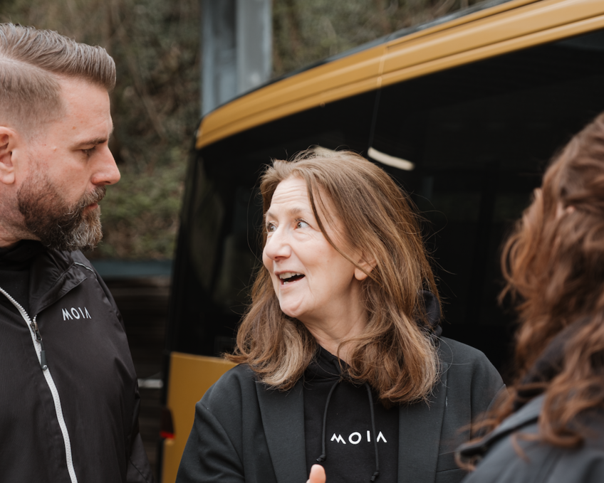 Three people wearing MOIA-branded clothing stand beside a MOIA vehicle and talk during a safety driver training session.