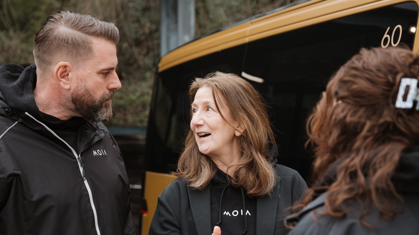Three people wearing MOIA-branded clothing stand beside a MOIA vehicle and talk during a safety driver training session.