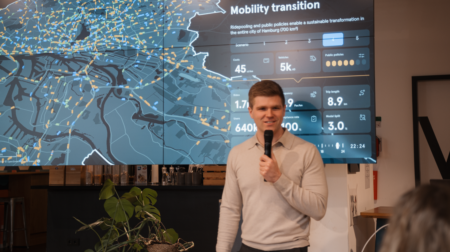A man holds a microphone and presents in front of a large screen displaying a city map and mobility data dashboard, illustrating traffic flows and performance metrics.