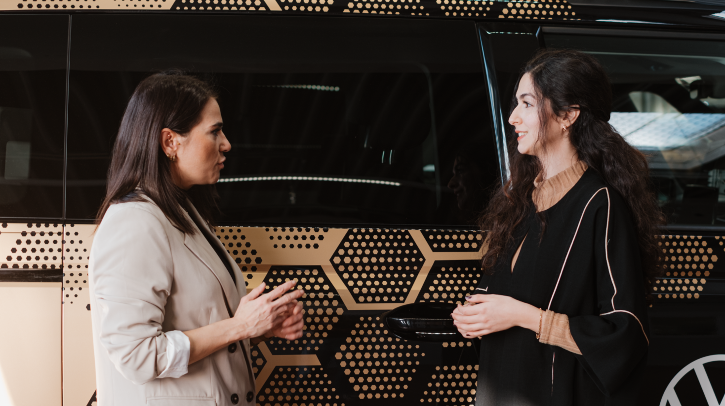 Two women stand facing each other and talk beside a vehicle with a patterned exterior, suggesting a discussion about mobility or automotive topics.