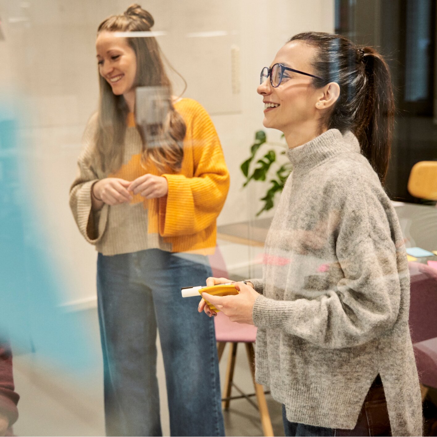Three colleagues stand in a modern office near a glass wall with sticky notes, smiling and discussing ideas; one holds a marker while the others listen and exchange ideas.