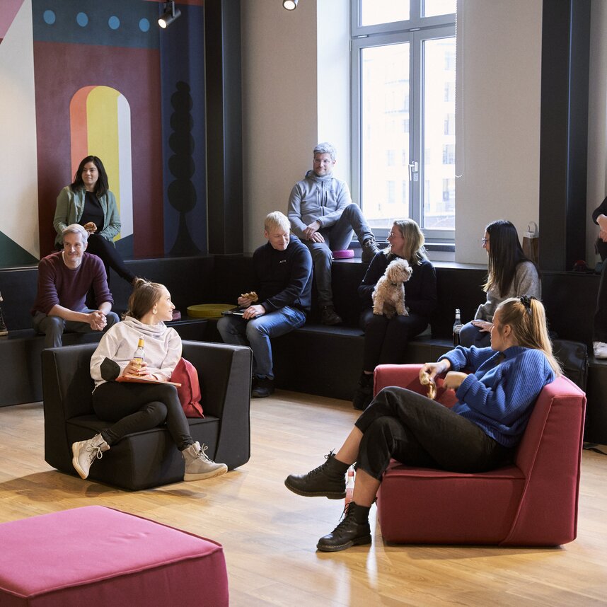 A group of people sitting on tiered seating and chairs in a modern office lounge, chatting and relaxing together; one person is holding a small dog, while others use laptops or talk in small groups near a large window and colorful wall mural.