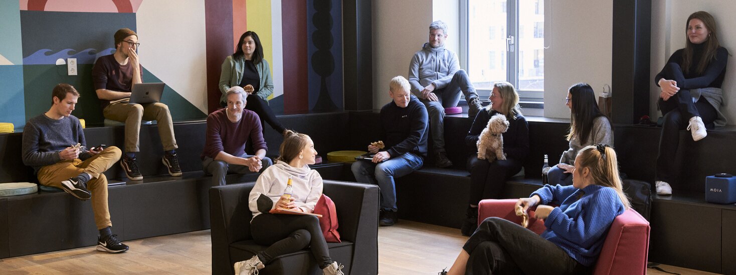 A group of people sitting on tiered seating and chairs in a modern office lounge, chatting and relaxing together; one person is holding a small dog, while others use laptops or talk in small groups near a large window and colorful wall mural.