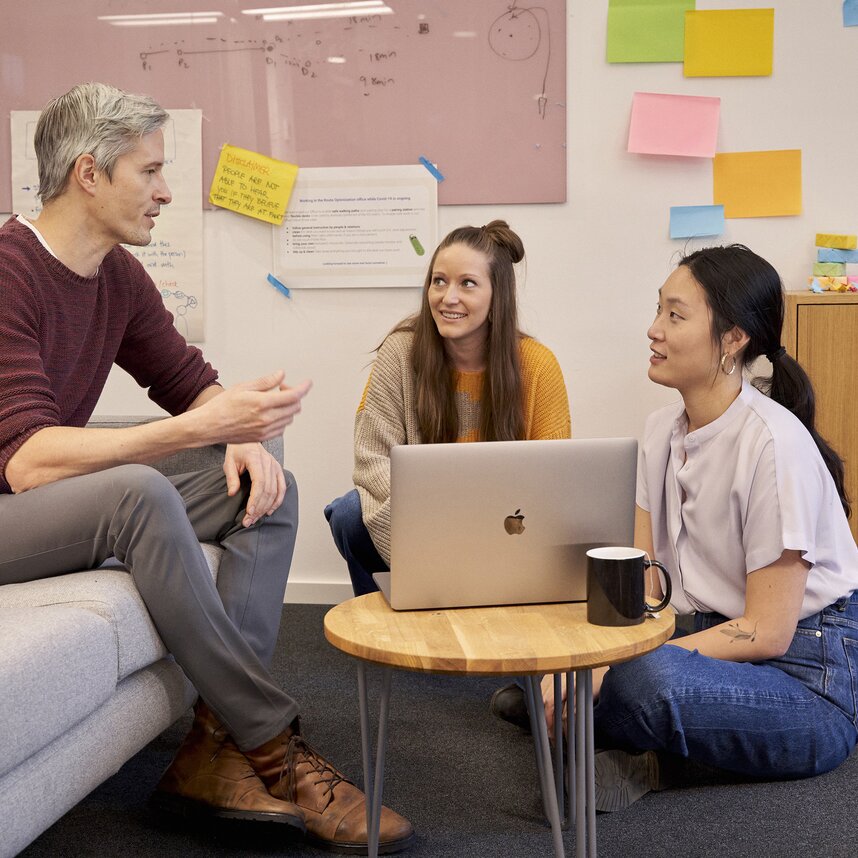 Three colleagues collaborate in a modern office, discussing ideas around a laptop with sticky notes and a whiteboard in the background.